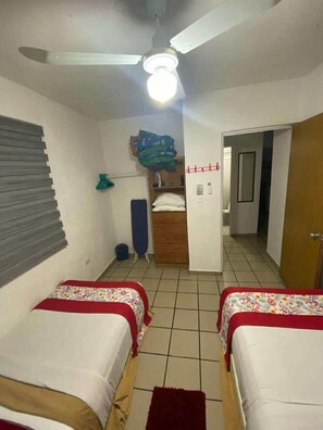 Una habitación con dos camas, un ventilador de techo, una ventana con persianas, una puerta y suelo de baldosas.