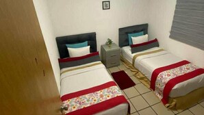 Ein Hotelzimmer mit zwei Betten, einem Deckenventilator, einem kleinen Nachttisch mit Pflanze und einem Fenster mit Jalousien.