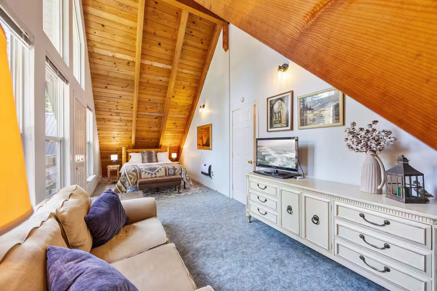A cozy attic bedroom with a sloped ceiling, a bed with a patterned blanket, a TV on a dresser, and a vase with flowers.