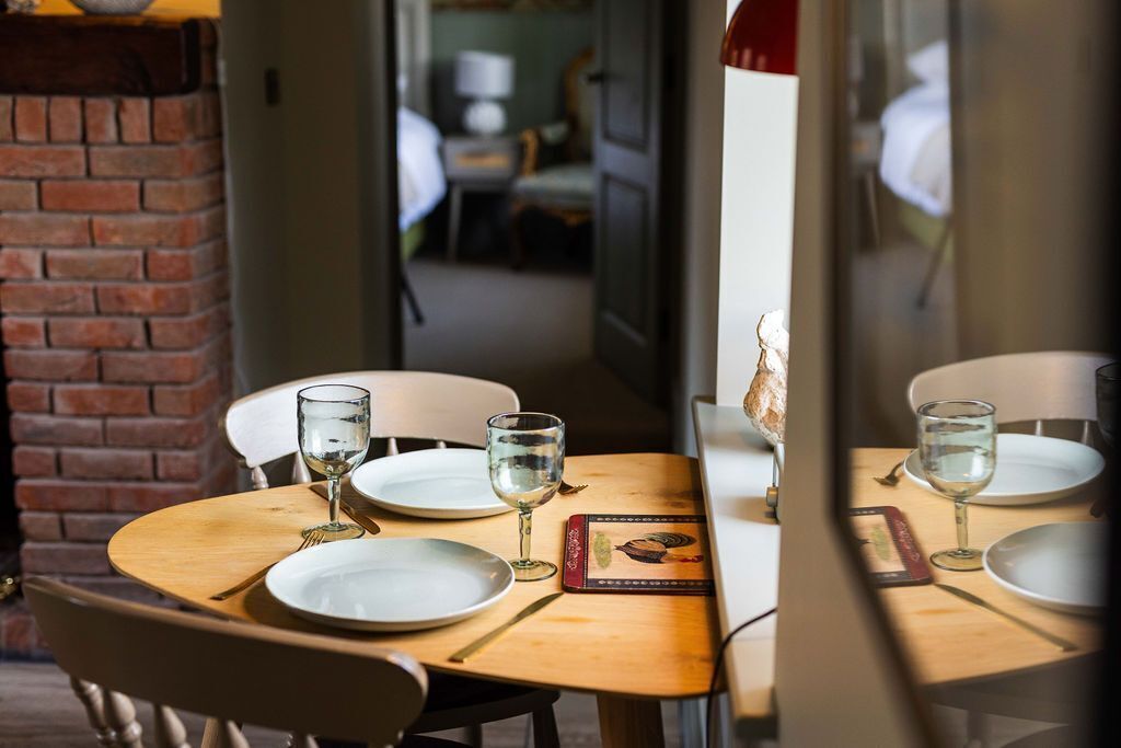 Una mesa de comedor servida con platos, vasos y cubiertos, con una pared de ladrillo y una puerta que da a otra habitación.