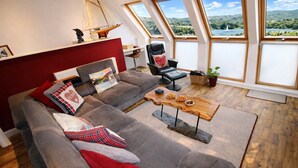 A modern loft-style living room with a large gray sofa, a wooden coffee table, and a view of a lake through skylights. - Romantic cottage with lake view in Bowness on Windermere (Bowness on Windermere)