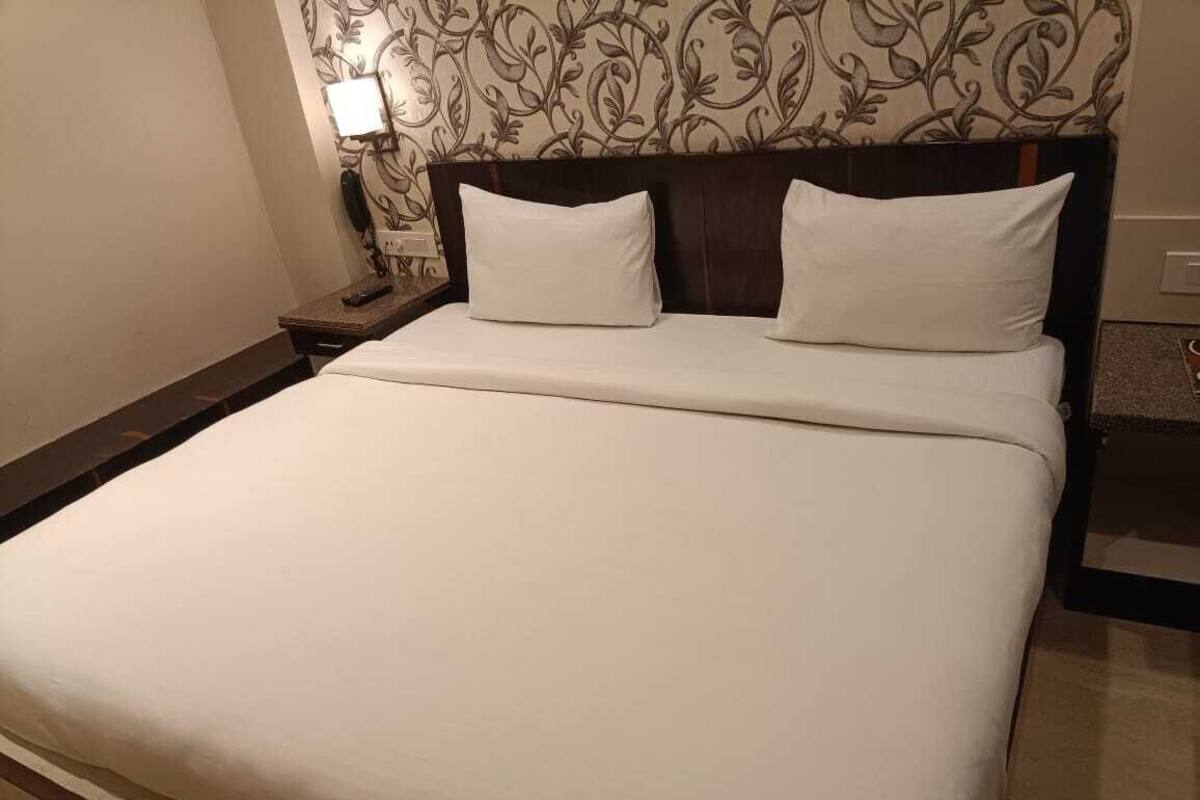 Quarto de hotel com cama grande, duas almofadas, mesa de cabeceira e luminária, além de papel de parede estampado.