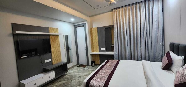 A modern hotel room with a large bed, a flat-screen TV, a vanity mirror, and a large window with curtains.