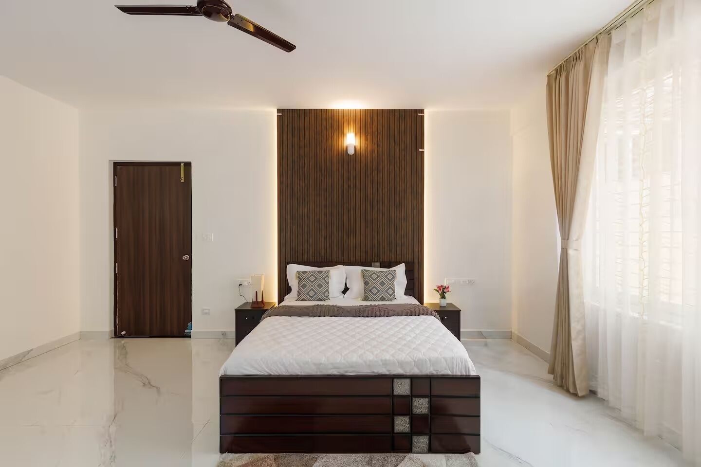 A bedroom with a bed, a nightstand, a ceiling fan, a wooden panel wall, and a door.