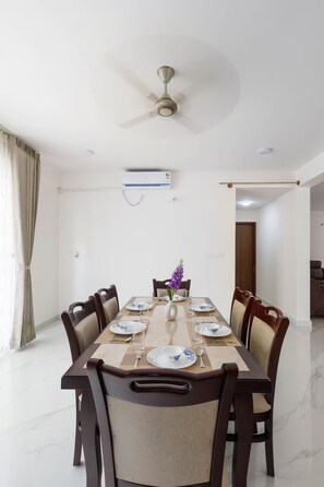 Une salle à manger avec une longue table dressée pour six personnes, entourée de chaises, un ventilateur de plafond et un appareil de climatisation.
