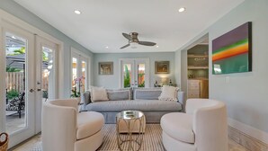 A modern living room with a grey sofa, two white armchairs, a glass coffee table, and a ceiling fan. - Pass A Grille (St. Pete Beach)