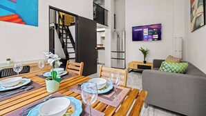 En spiseplads med et træbord dækket til fire personer, en trappe, et køleskab og et fladskærms-TV der viser streamingtjenester.
