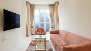 A modern living room with a flat-screen TV, a pink sofa, a small table with a vase, and a view of a cityscape through large windows. - Casa Opal by Red Olive1bhkleela Residency (Bengaluru)