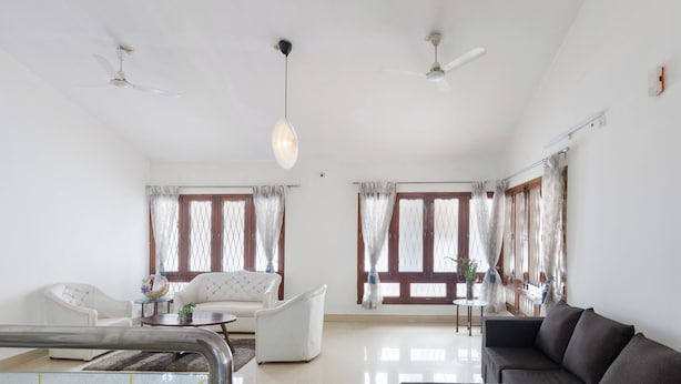 A spacious living room with a sofa, armchairs, a coffee table, and a dining table. There are two ceiling fans, a hanging light fixture, and large windows with curtains. - Casa Promenade by Red Olive 3bhk Frazer Town (Bengaluru)