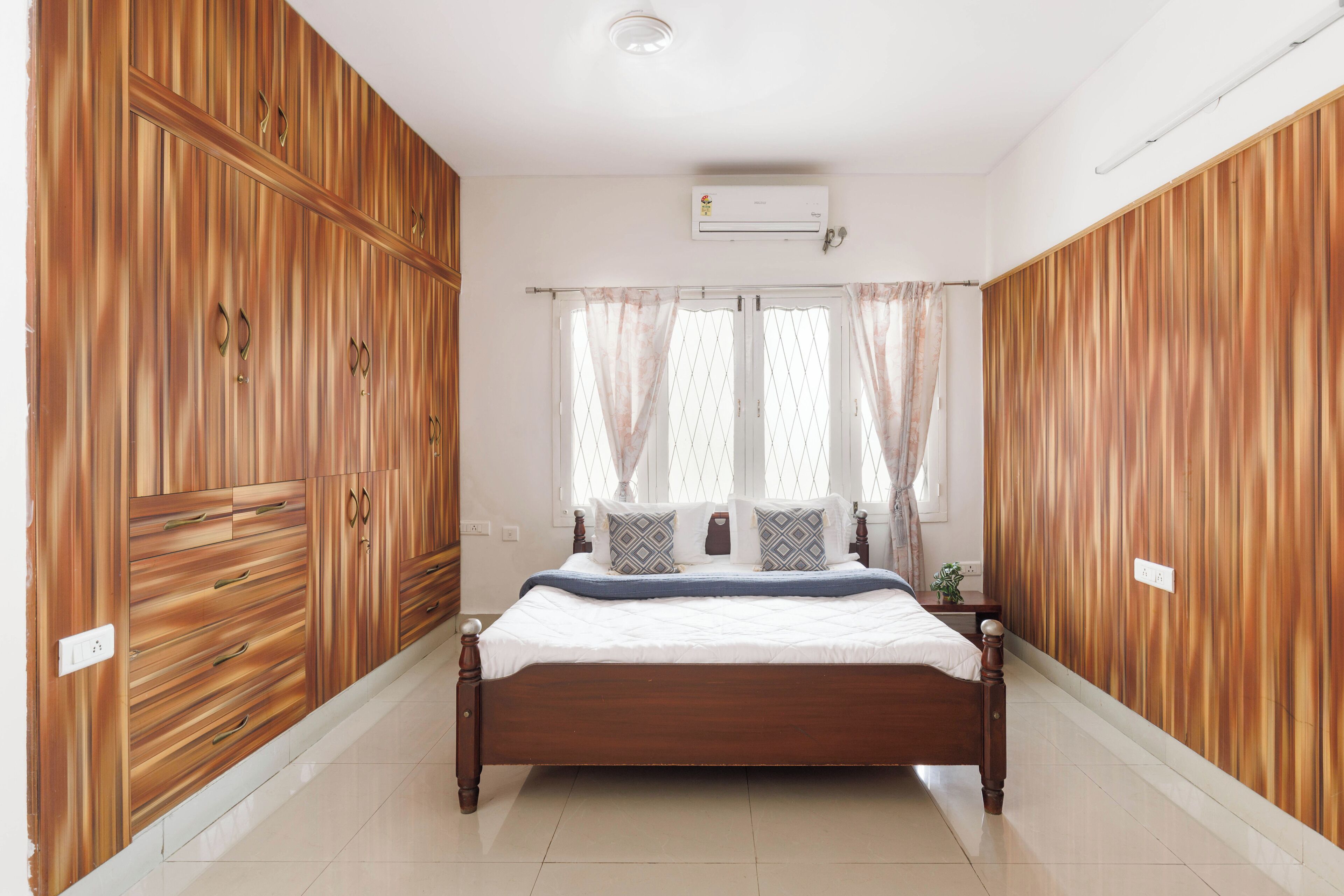 A bedroom with a wooden wardrobe, a bed with a white mattress and blue bedding, and a window with sheer curtains.