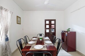 Uma sala de jantar com uma mesa de madeira posta para quatro pessoas, rodeada por cadeiras, um ventilador de teto e uma geladeira.