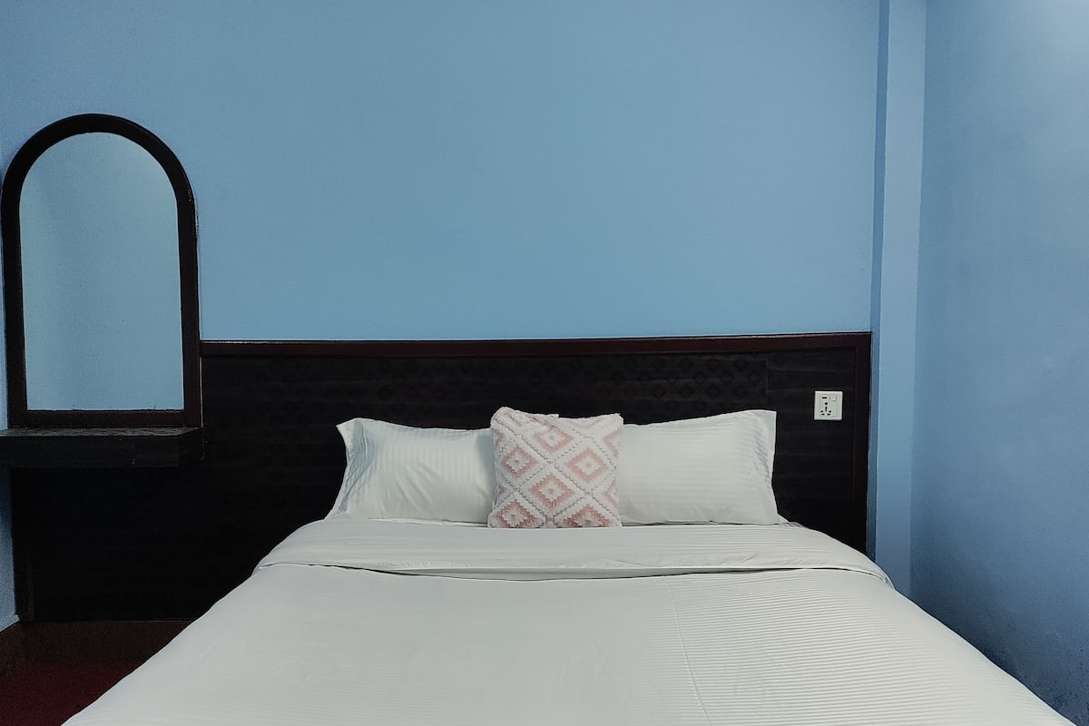A neatly made bed with white bedding and two pillows, a dark headboard, and a wall-mounted mirror.