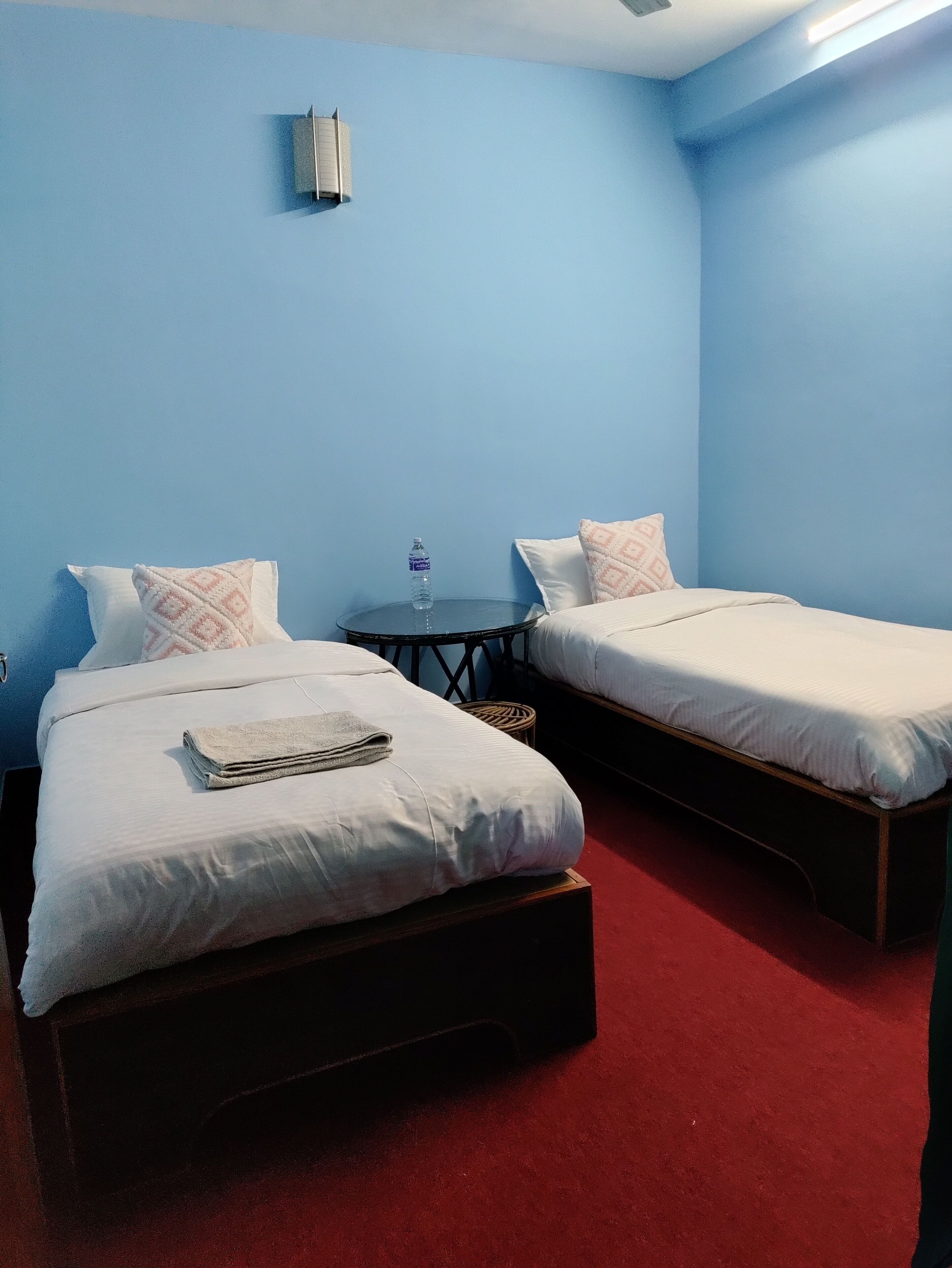 A hotel room with two single beds, a small table with a water bottle, and a wall-mounted air conditioner.