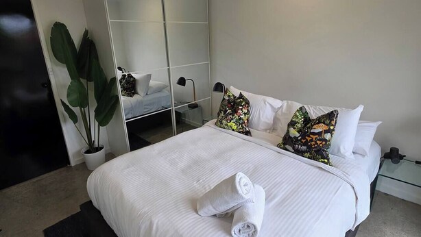 A neatly made bed with white linens and rolled towels, a large mirror, and a potted plant in a modern hotel room. - A Sporting Spot in Warm Sunshine (Sunshine North)