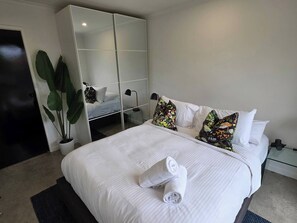 Un lit bien fait, avec du linge de lit blanc et des serviettes pliées, un grand miroir et une plante en pot, dans une chambre d’hôtel moderne.