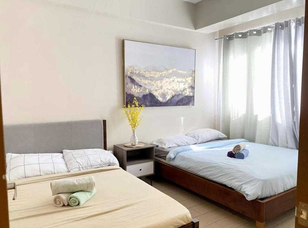 Habitación de hotel con dos camas, una mesita de noche con un jarrón de flores, un cuadro en la pared y una ventana con cortinas.