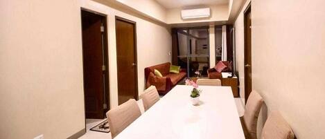 A modern living room with a large white dining table, comfortable seating, and a sliding glass door leading to a balcony.
