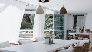 Comedor con una mesa grande, sillas de mimbre, lámparas colgantes y vistas a exuberante vegetación a través de amplios ventanales.