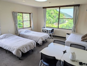 Une chambre d’hôtel avec deux lits, une petite table, un canapé et de grandes fenêtres donnant sur un paysage verdoyant.