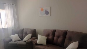 A living room with a brown sofa and a brown armchair, both with white cushions. - Aluguel season (Guarapari)