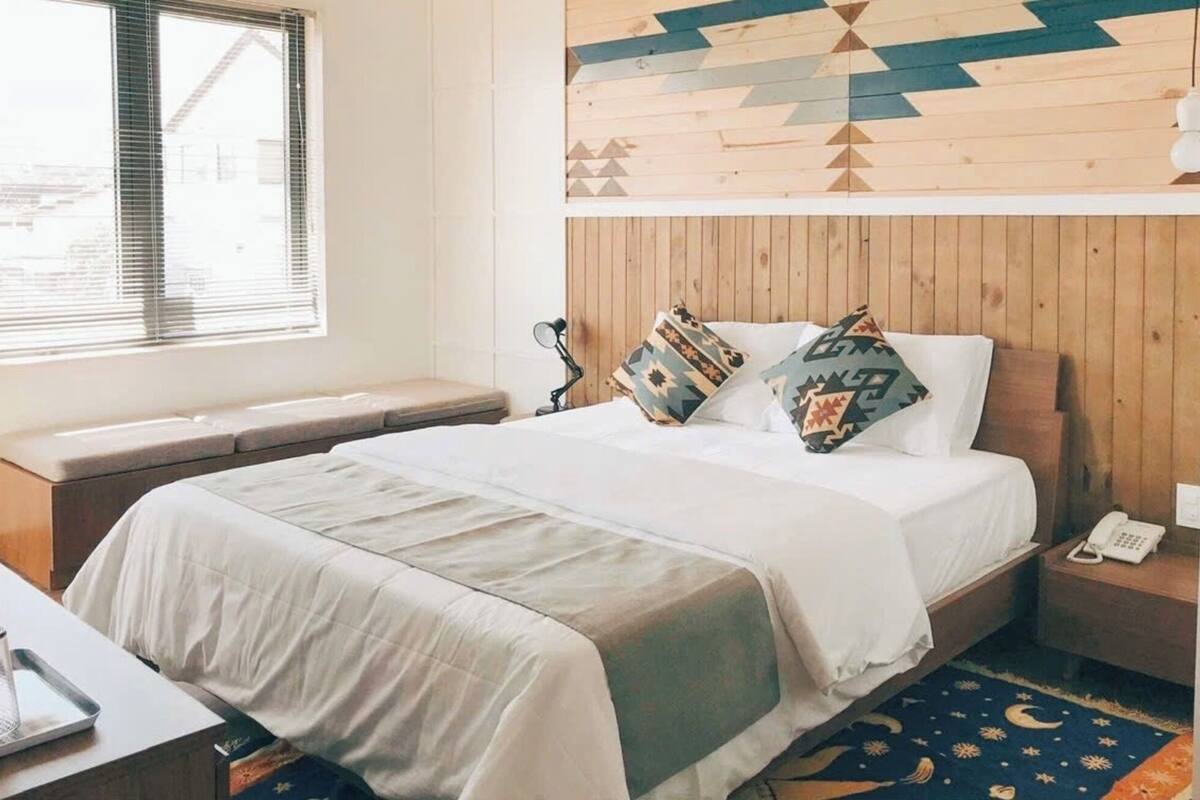 Un dormitorio con cabecera de madera, una cama con almohadillas estampadas, una mesita de noche con lámpara y una ventana con persianas.