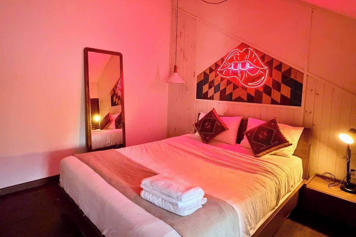 Una habitación de hotel con una cama, un espejo, un letrero luminoso con forma de labios y una mesita de noche con una lámpara.
