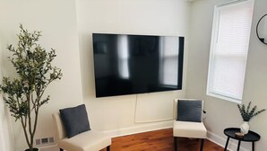 Een moderne woonkamer met een flatscreen televisie, twee witte banken met donkere kussens, een houten vloer en een grote potplant.