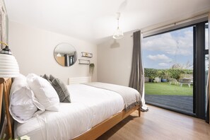 A bedroom with a large bed, a round mirror, a ceiling light, and a view of the garden through a sliding glass door. - Cloud 10 St Ives - Sea Views - Free parking & WiFi - Pets Welcome (St Ives)