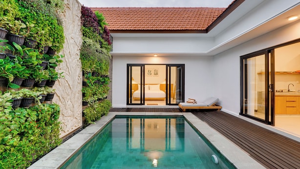 A modern outdoor pool area with a wooden deck, a stone wall with greenery, and a glimpse of a bedroom with a bed and a nightstand. - Bhatara Villa Sanur (Denpasar)