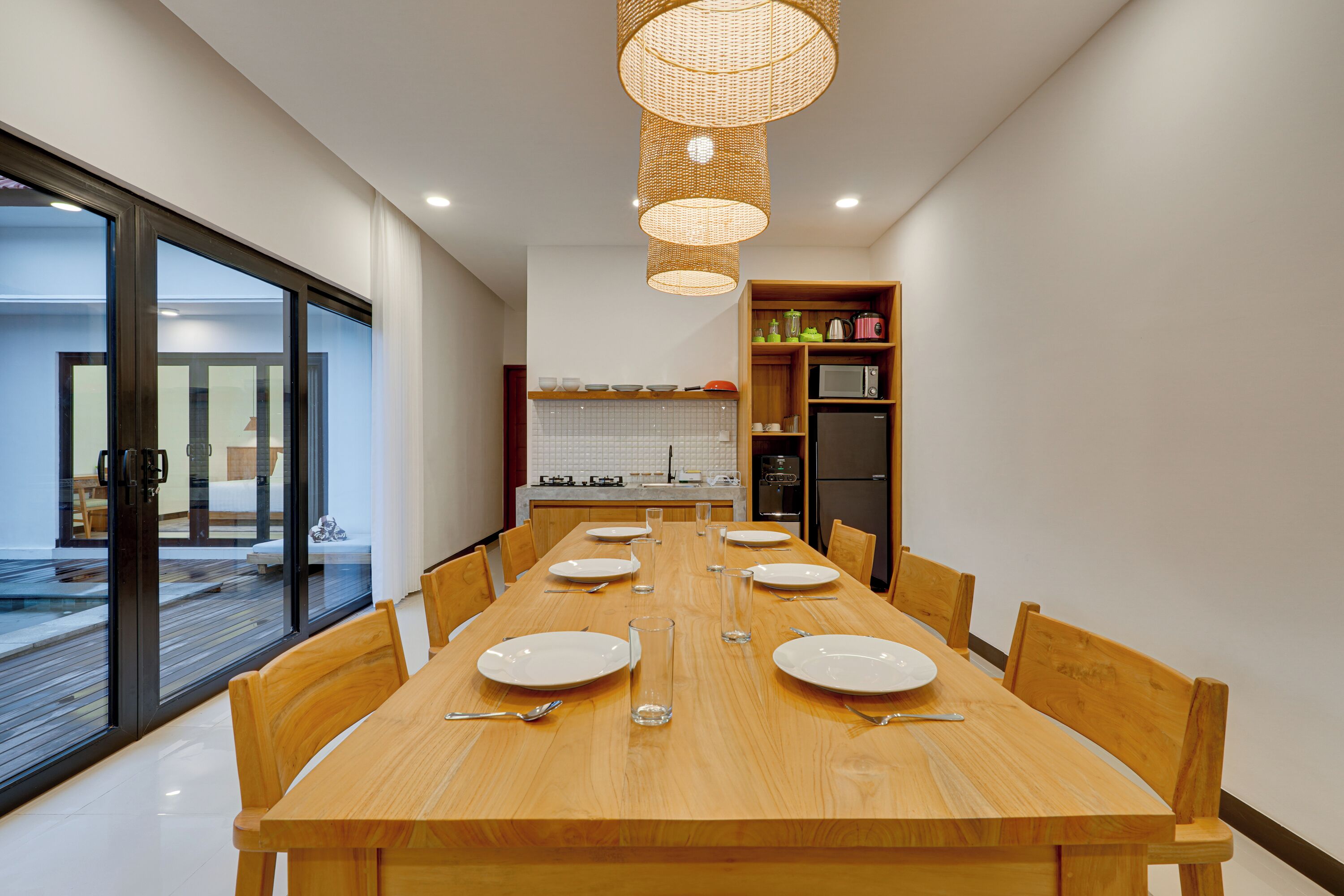 A modern dining area with a long wooden table set for six, surrounded by wooden chairs. A large window provides a view of the outdoor deck.