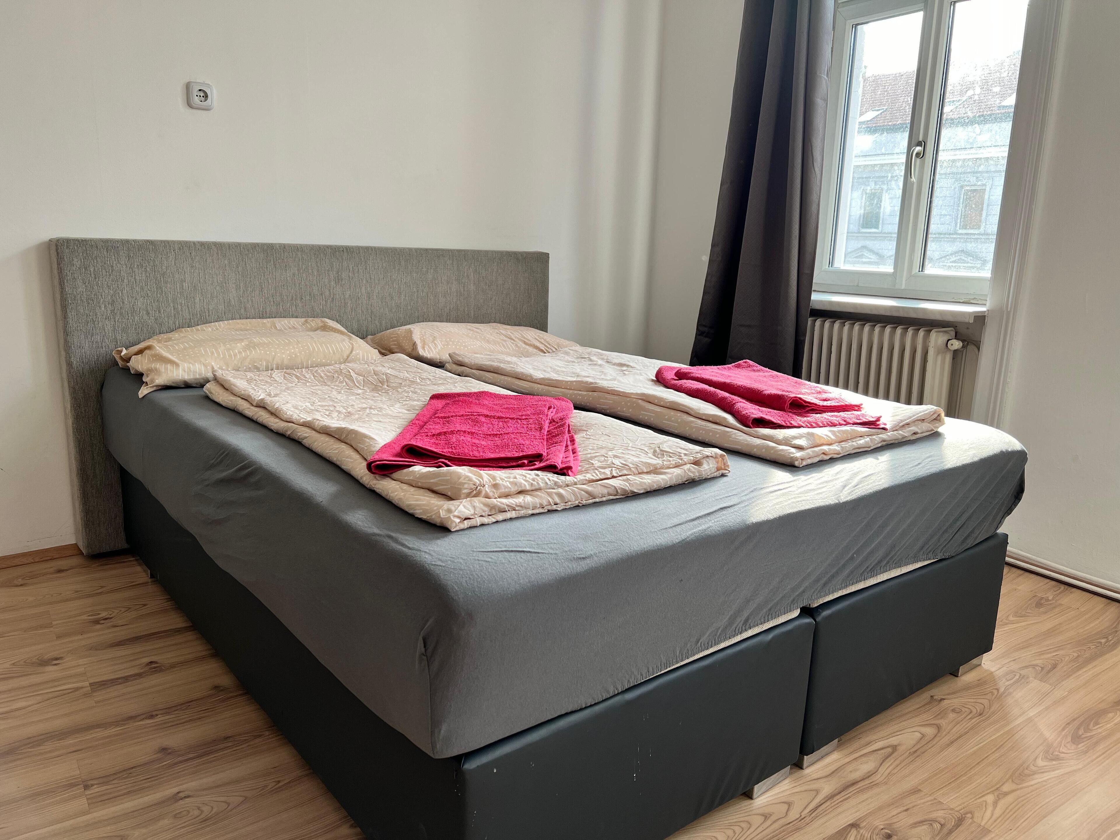 Une chambre moderne avec un lit recouvert d’un tissu gris, deux oreillers et une pile de serviettes pliées.