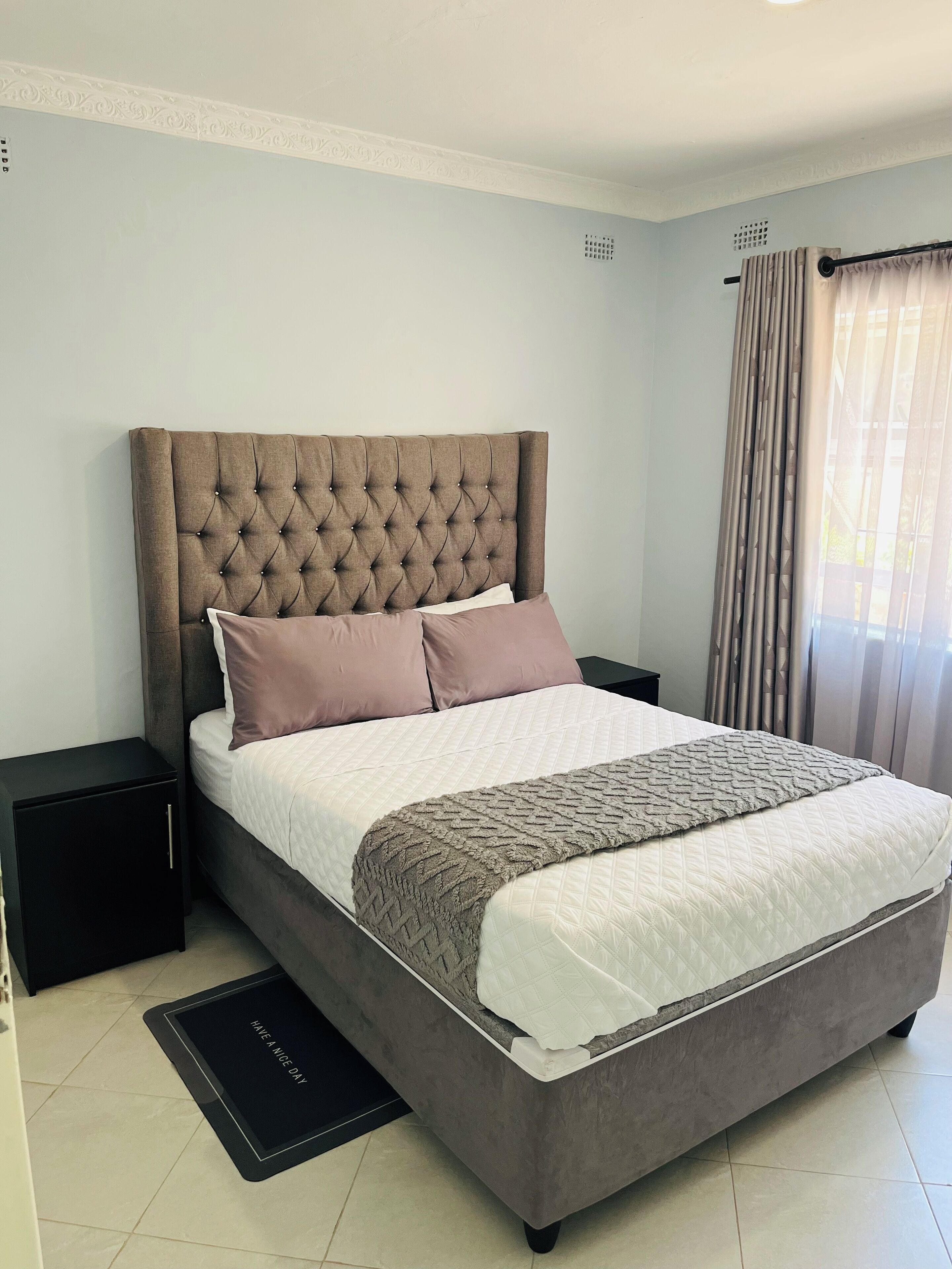 Un dormitorio con cabecera capitonada, cama con ropa blanca y marrón, mesita de noche y ventana con cortinas.