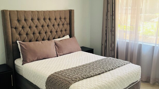 A bedroom with a large bed, a nightstand, a black doormat, and a window with curtains.