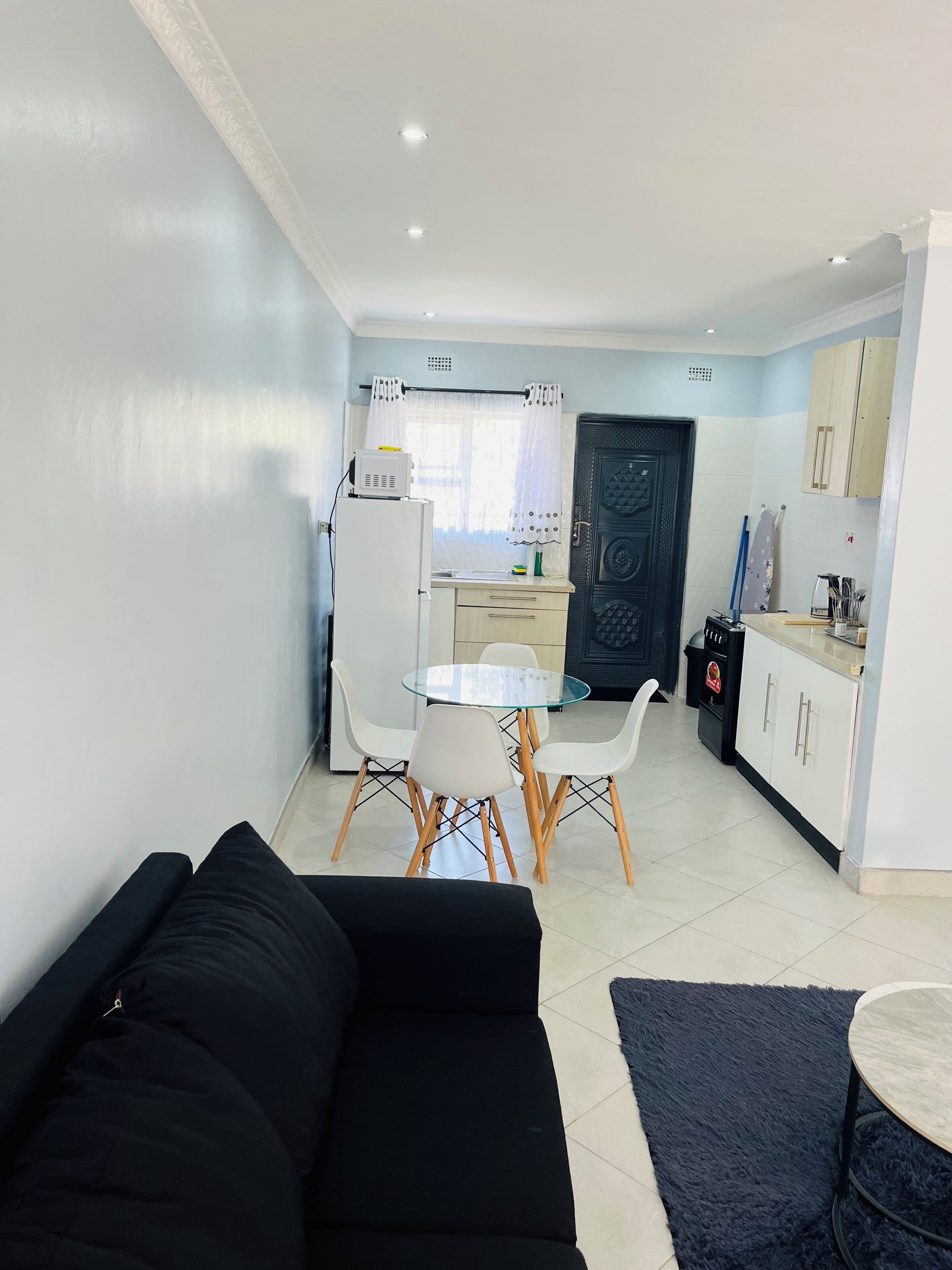 Una sala de estar moderna con un sofá negro, una mesa de comedor de vidrio con sillas blancas, y una zona de cocina con un refrigerador, estufa y armarios.