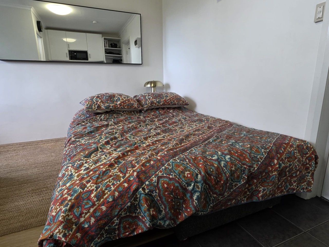 A bed with a patterned bedspread, a bedside lamp, and a mirror on the wall.