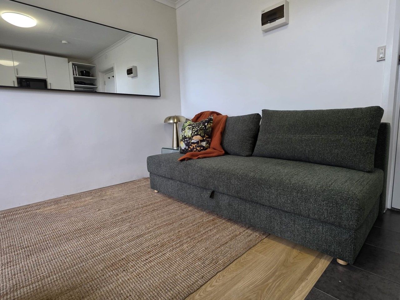 A room with a grey sofa, a patterned rug, a mirror, and a small shelf.