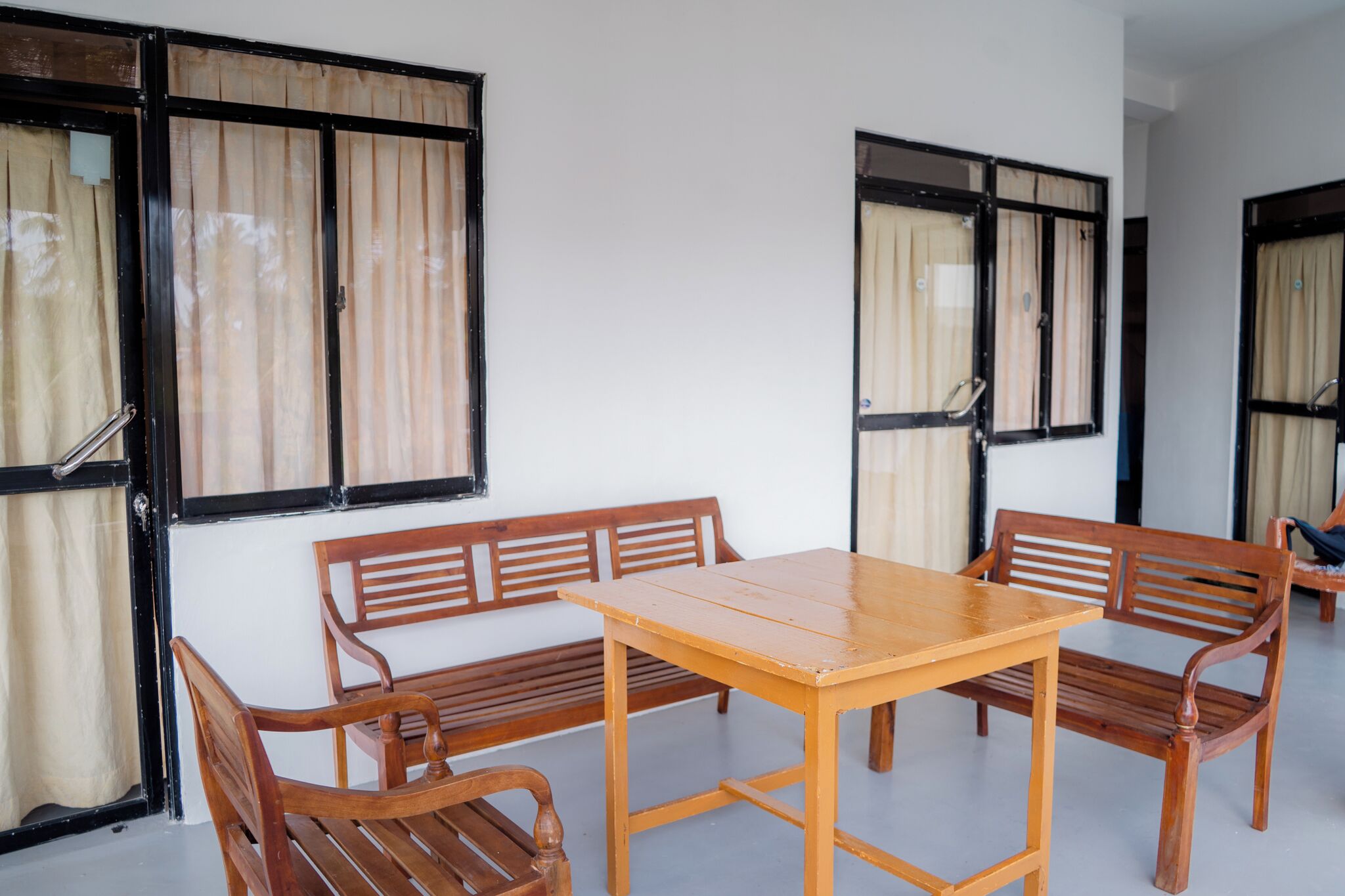 Una habitación con una mesa de madera y bancos, ventanas amplias con cortinas y una pared blanca.