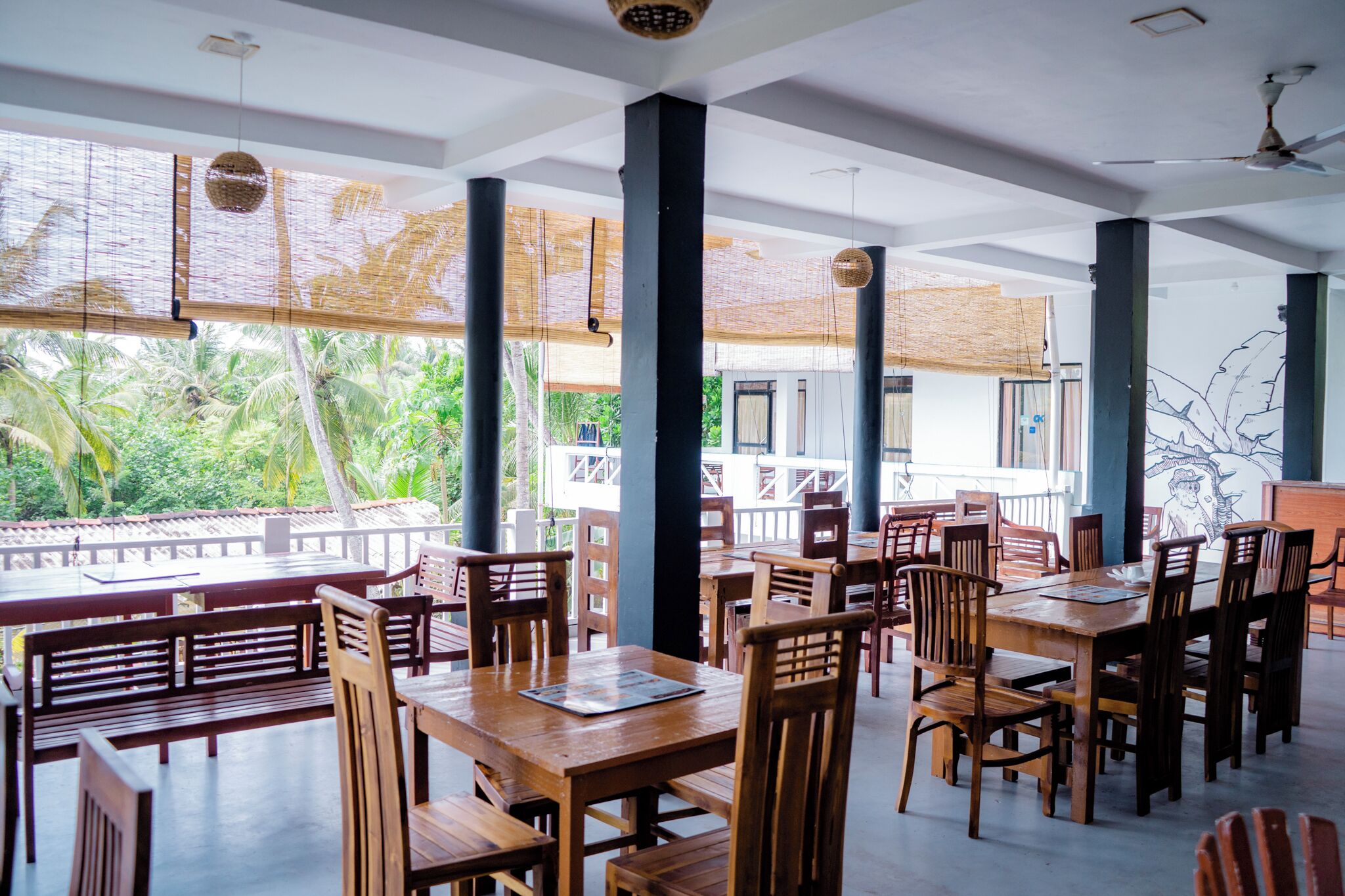 A restaurant with wooden tables and chairs, large windows with a view of palm trees, and a ceiling fan.