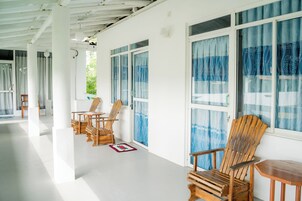Een overdekt terras met houten stoelen en tafels, grote ramen met blauwe gordijnen, en een witte plafondconstructie.