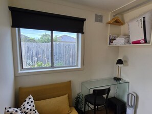 A small, tidy room with a bed, a desk, and a chair. There is a window with a view of a fence and trees. - A Sweet Studio for One (Seaholme)