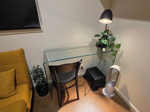 A compact workspace with a glass desk, a chair, a small heater, a potted plant, and a yellow couch. - A Sweet Studio for One (Seaholme)