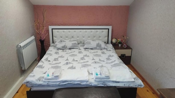 Uma cama bem arrumada com colcha estampada, uma mesa de cabeceira com um globo e flores, e um radiador na parede.