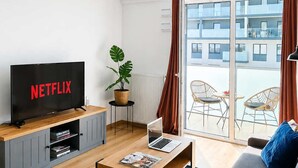 Een moderne woonkamer met een flatscreen televisie, een laptop, een salontafel, een bank met kussens, een potplant en uitzicht op een gebouw door een glazen deur.