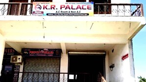 Un edificio de dos pisos con un cartel que dice K.R. Palace A.C Room & Non A.C. Abajo, hay un cartel de agente de propiedades y un anuncio de Sprite.
