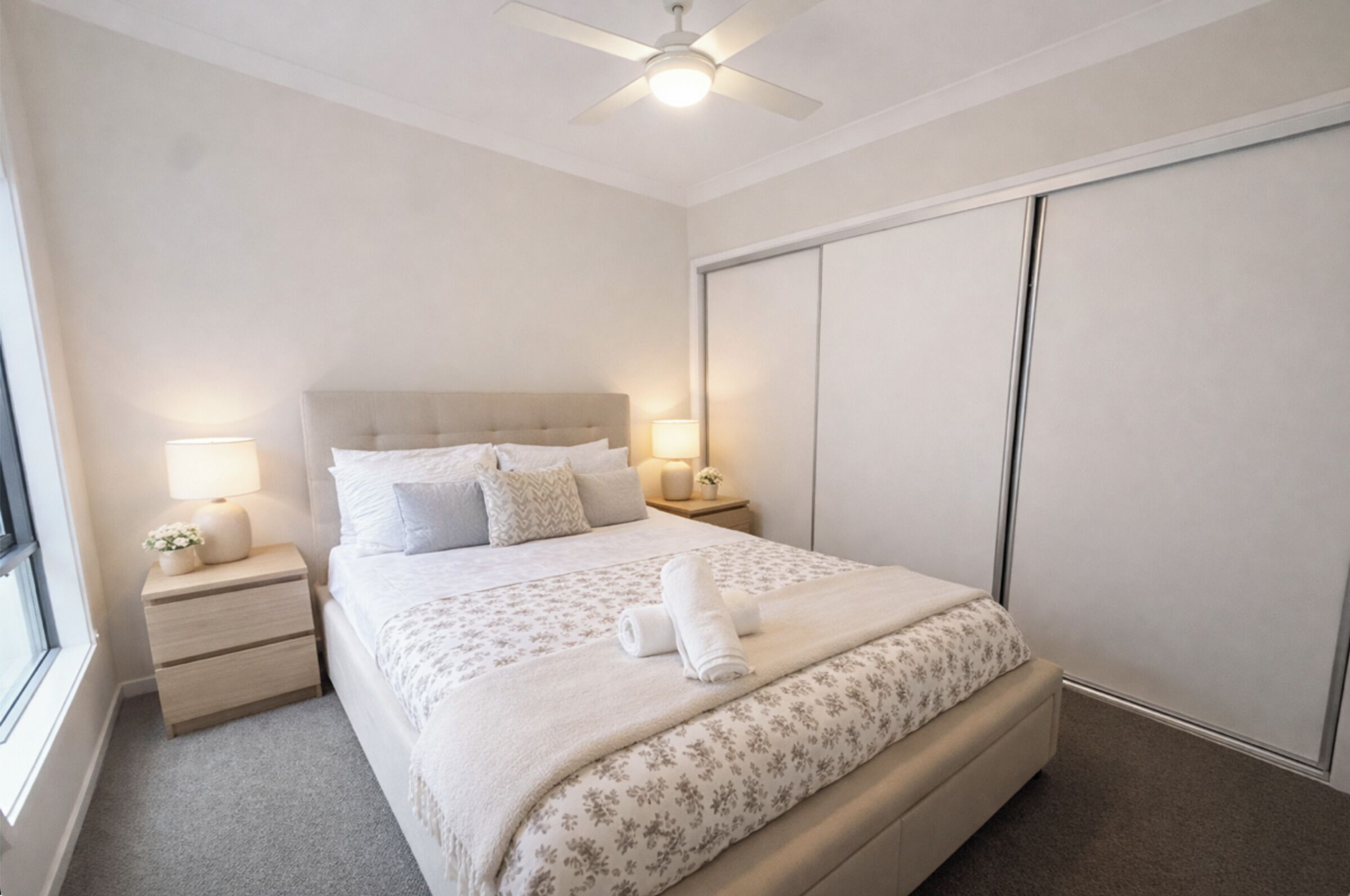 Ein ordentlich bezogenes Bett mit floralem Überwurf, zwei Nachttischlampen, einem Deckenventilator und einem großen Kleiderschrank.