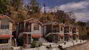 A row of modern, multi-story cabins with stone exteriors and wooden accents, set against a mountainous backdrop. - Sparsh Himalaya Shivir (Rishikesh)