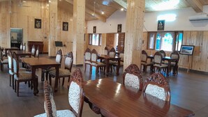 A spacious dining area with wooden tables and chairs, framed artwork on the walls, and large windows allowing natural light. - Sparsh Himalaya Shivir (Rishikesh)