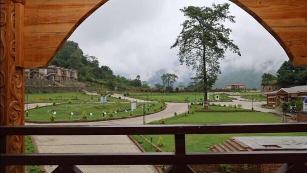 A view through a wooden archway of a landscaped area with pathways, a central tree, and buildings in the background. - Sparsh Himalaya Shivir (Rishikesh)