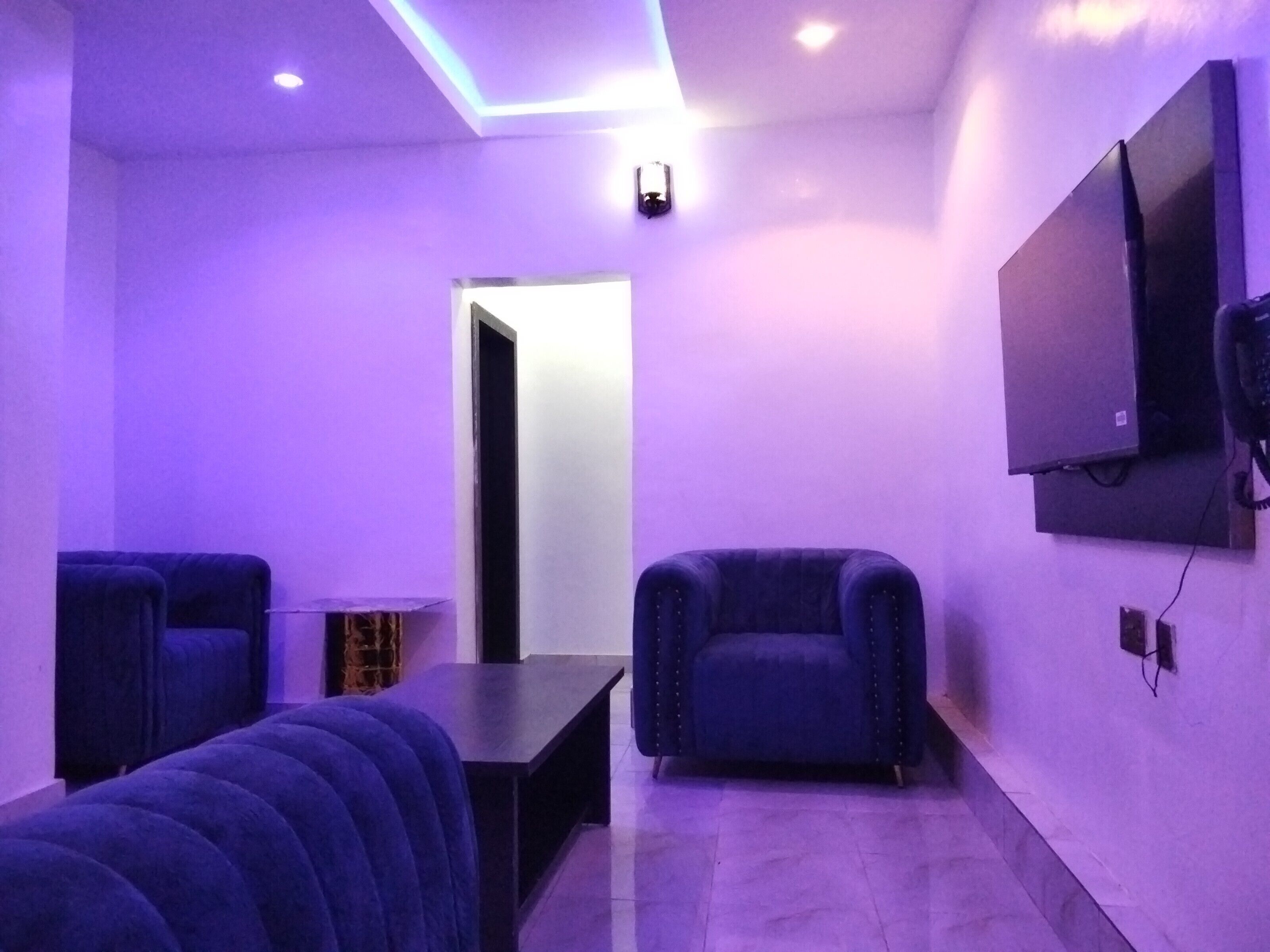 Una habitación con un ambiente iluminado en morado, que incluye un sofá, una mesita y un televisor montado en la pared.