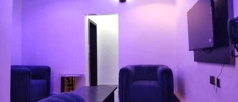 A room with a purple-lit ambiance, featuring a sofa, a small table, and a TV mounted on the wall.